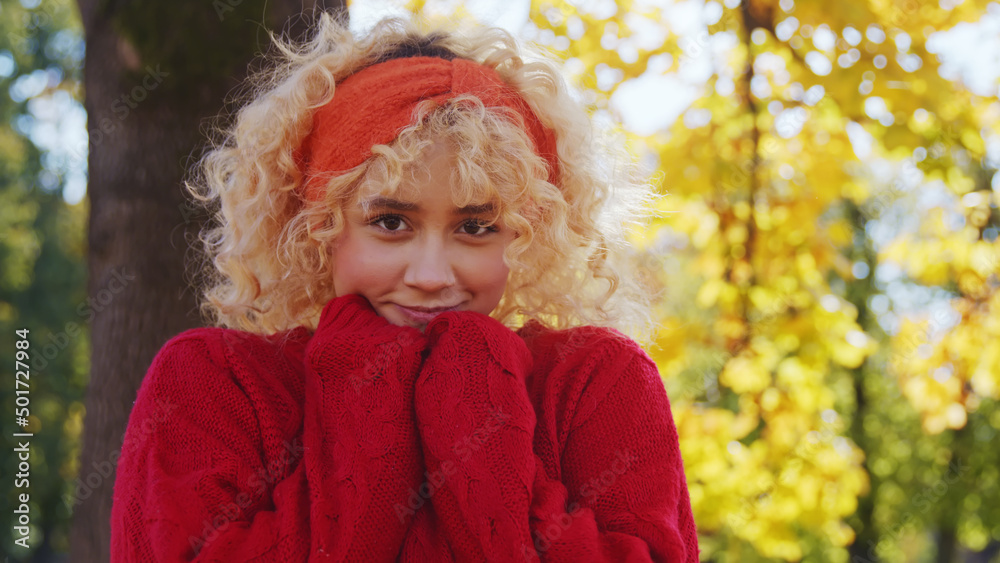 Portrait of a beautiful caucasian European blonde curly-haired girl smiling in a shy way and looking at camera with her palms hidden in the sleeves of her red sweater. Autumn scenery. High quality Portrait of a beautiful caucasian European blonde curly-haired girl smiling in a shy way and looking at camera with her palms hidden in the sleeves of her red sweater. Autumn scenery. High quality