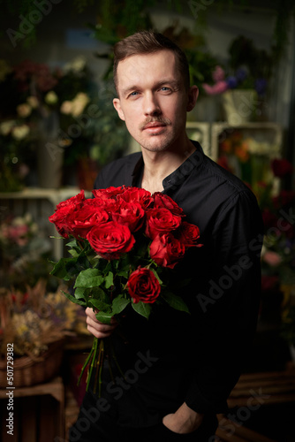 Romantic, dating or love concept. Stylish sexy macho man with mustache holding red roses bouquet in hand, looking at camera wearing black shirt in flower store with dark background. High quality photo