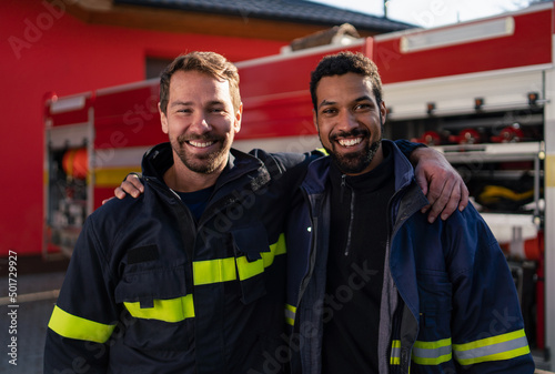 Happy firefighters crew with fire station and truck in background looking at camera.