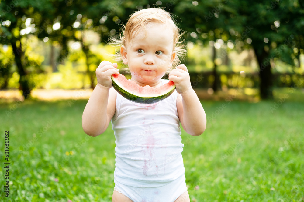 funny smiling kid boy in white bodysuit eating watermelon at green lawn funny smiling kid boy in white bodysuit eating watermelon at green lawn