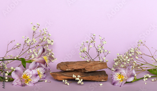 Empty pedestal made of natural brown stone for presentation of cosmetic products and delicate flowers of gypsophila and alstroermeria on violet  background. Advertising of spring rejuvenating cosmetic