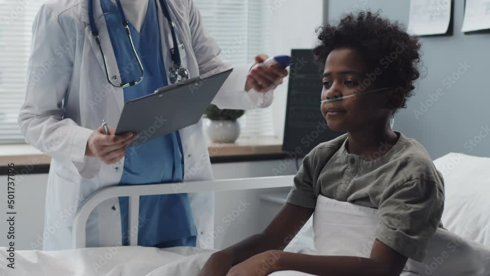 Locked down of African American boy with oxygen tube in nose lying in bed, then sitting up when cropped doctor coming and measuring his body temperature with contactless thermometer