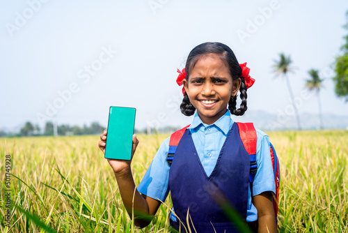 Happy smiling village girl kid showing green screen mobile phone while standing at paddy farmfield - concept of advertisement, app promotion and education photo