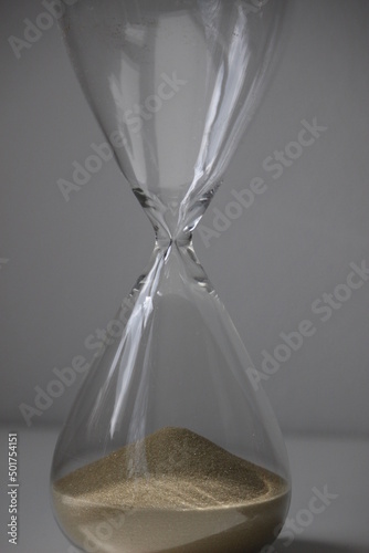 Glass Hourglass with most of it`s sand in the top section. The sand is beige and light and the object is in front of a white wall on top of a white table underground  photo