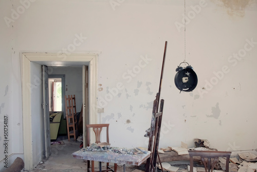 A room inside an abandoned house, used as a painter's studio: a canvas on the easel, many dried up colors on a table, a hanging clock, ruined books, everything is dirty and messy.
