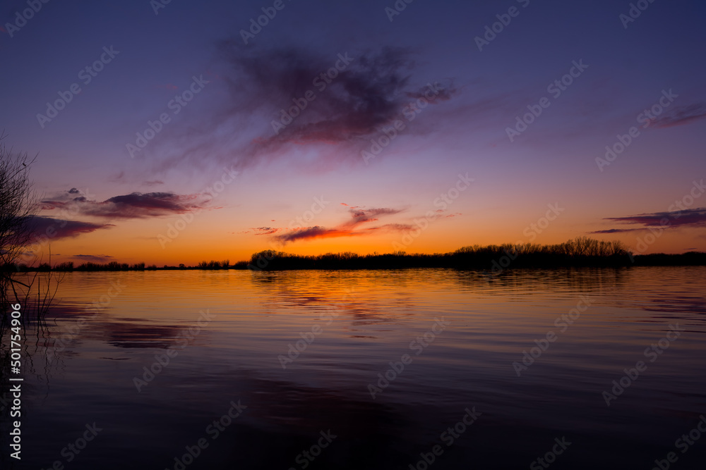Orange purple and violet sunset on river with dark colorful clouds in sky with trees reflection in water