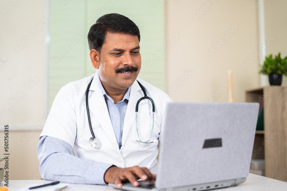 smiling doctor while busy using laptop at hospital - concept of online healthcare consultant, medicare and technology. smiling doctor while busy using laptop at hospital - concept of online healthcare consultant, medicare and technology.