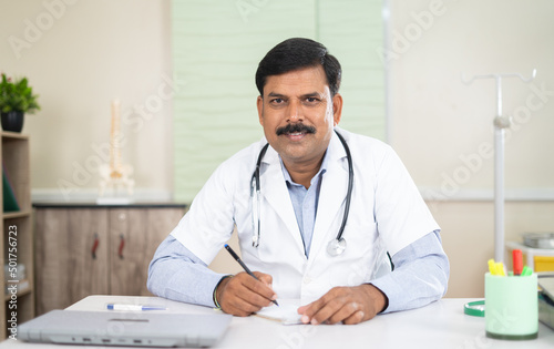 Doctor at clinic looking at camera while busy writing prescription at hospital - concept of professional occupation  happiness and healthcare or medical service
