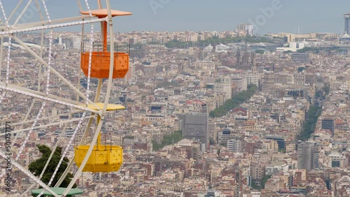 Empty gondolas of carousel wheel move against dense build city seen from height, fine telephoto perspective of Barcelona. Amusement ride at top of Tibidabo hill photo