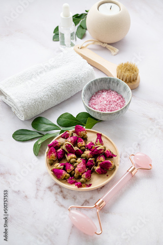 Organic sea salt for the body with dried rose flowers, candle, brush, towel and gua sha on a pink marble background. The concept of a natural spa product. Skin care