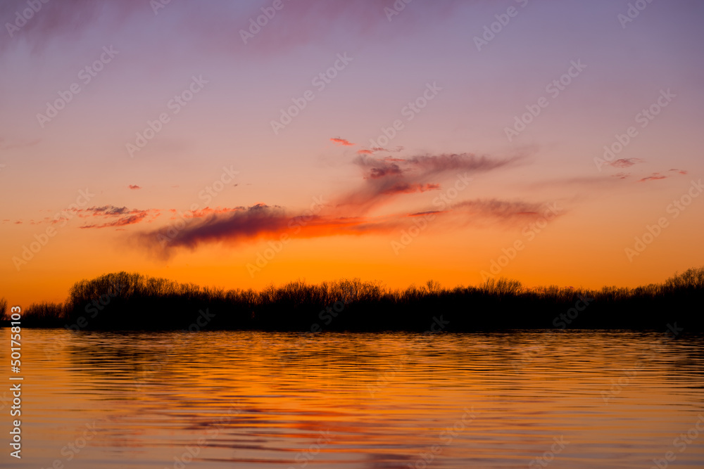 Orange purple and violet sunset on river with dark colorful clouds in sky with trees reflection in water 