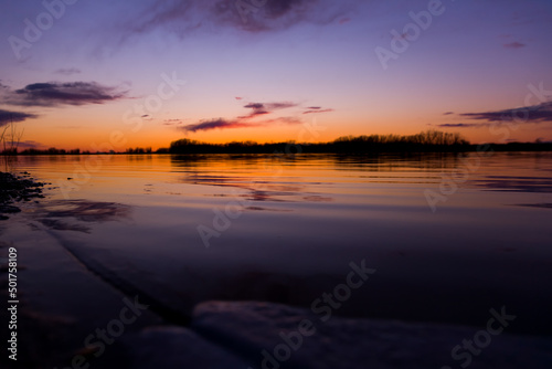 Orange purple and violet sunset on river with dark colorful clouds in sky with trees reflection in water