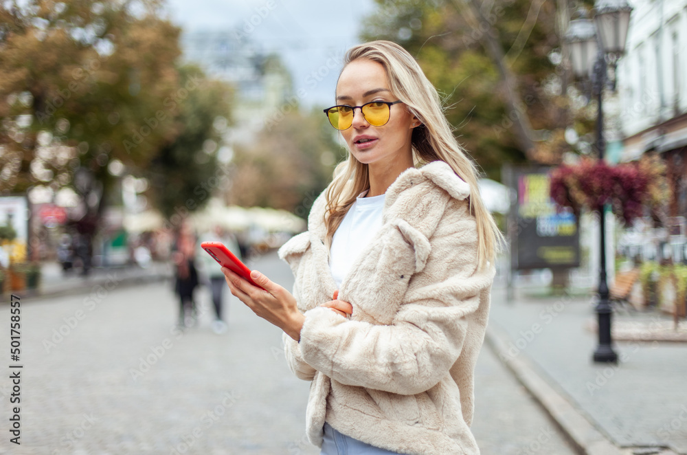 Obraz premium Stylish fashion woman in a fashionable fur coat and yellow glasses uses a smartphone in the city