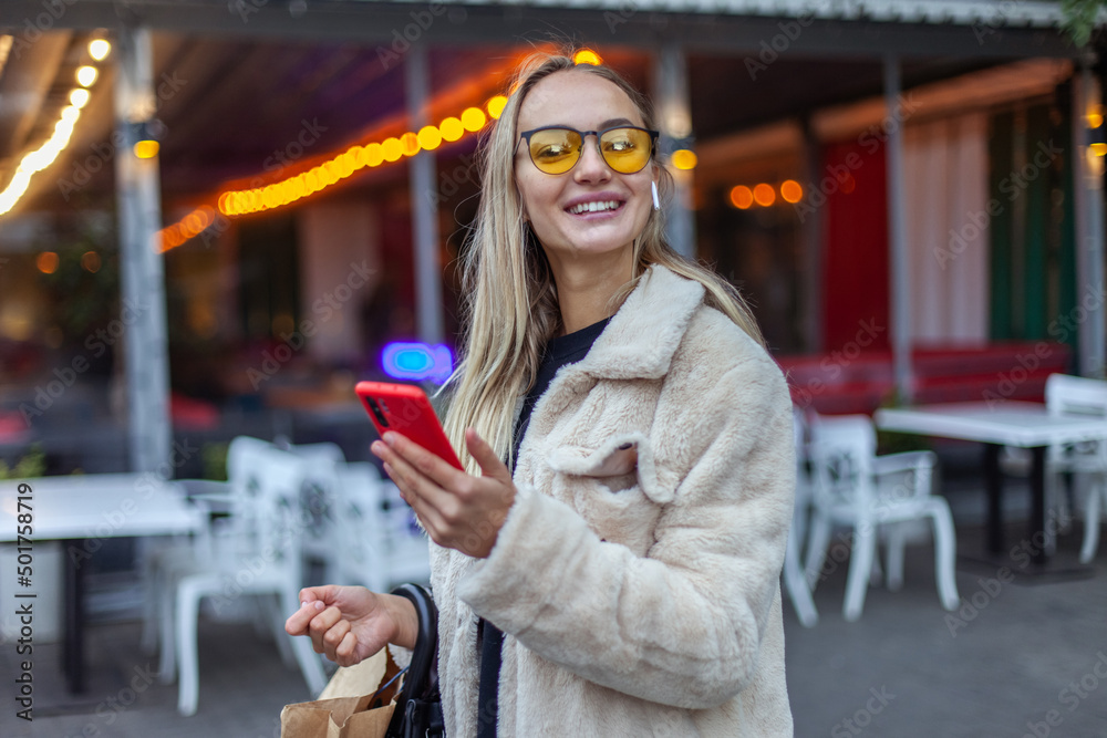 Young happy woman in yellow sunglasses and earphones using smartphone in evening city Young happy woman in yellow sunglasses and earphones using smartphone in evening city