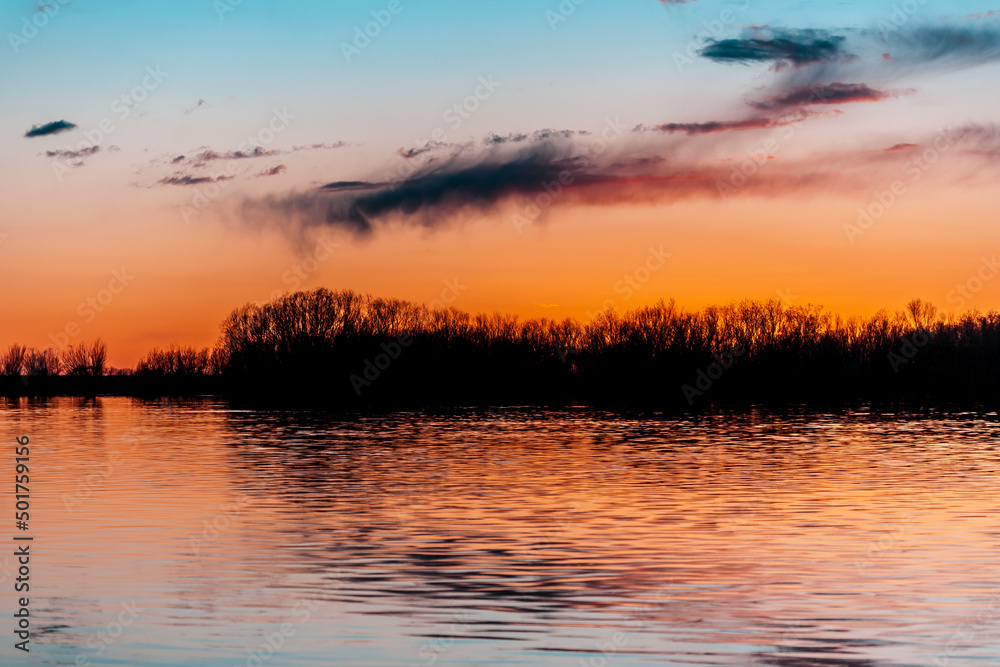 Orange purple and violet sunset on river with dark colorful clouds in sky with trees reflection in water 