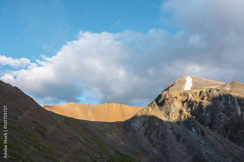 Scenic landscape with sunlit wide sharp mountain ridge between mountain tops at changeable weather. Colorful mountain scenery with large sharp rocks on ridge top in sunlight under clouds in blue sky.