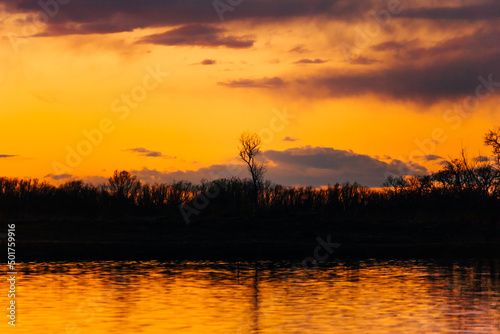 Orange purple and violet sunset on river with dark colorful clouds in sky with trees reflection in water 