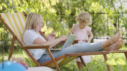Happy smiling family in green garden, sunny summer day. Mother with laptop and her little child girl have fun playing together. Daughter brings sunglasses to her mom, jumping joyfully. Mom kisses her