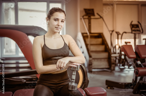 Slim young woman sitting on a bench with dumbbells in the gym
