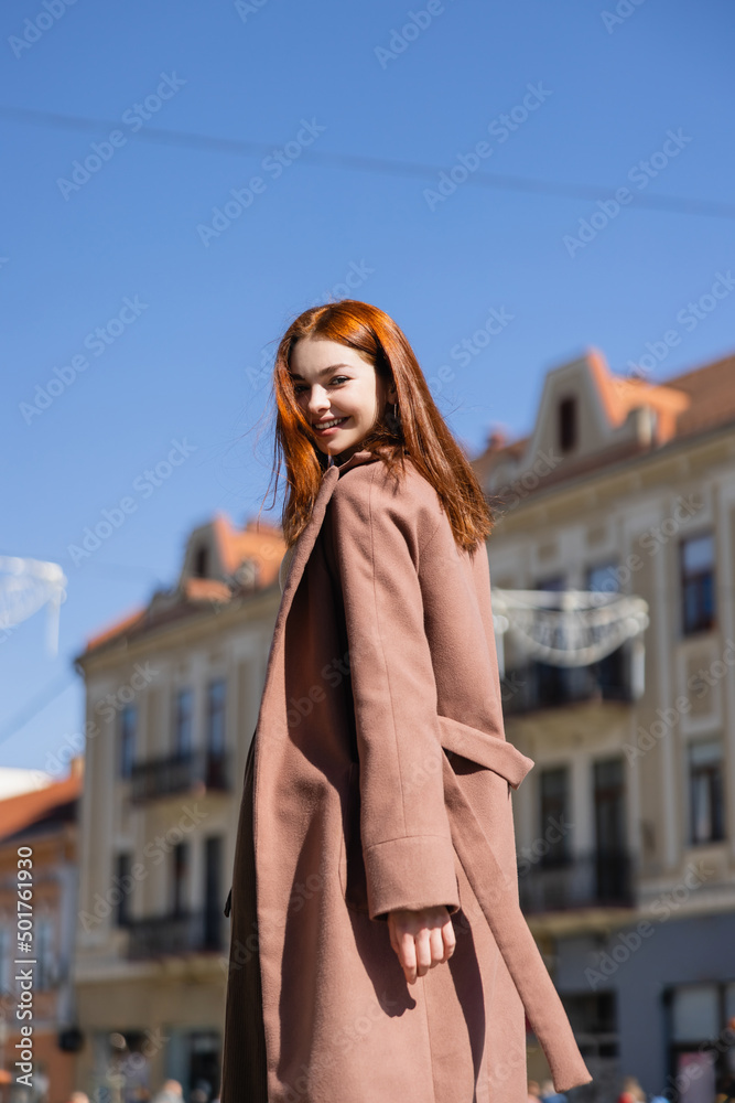 Fototapeta premium joyful redhead woman in coat on street of european city.