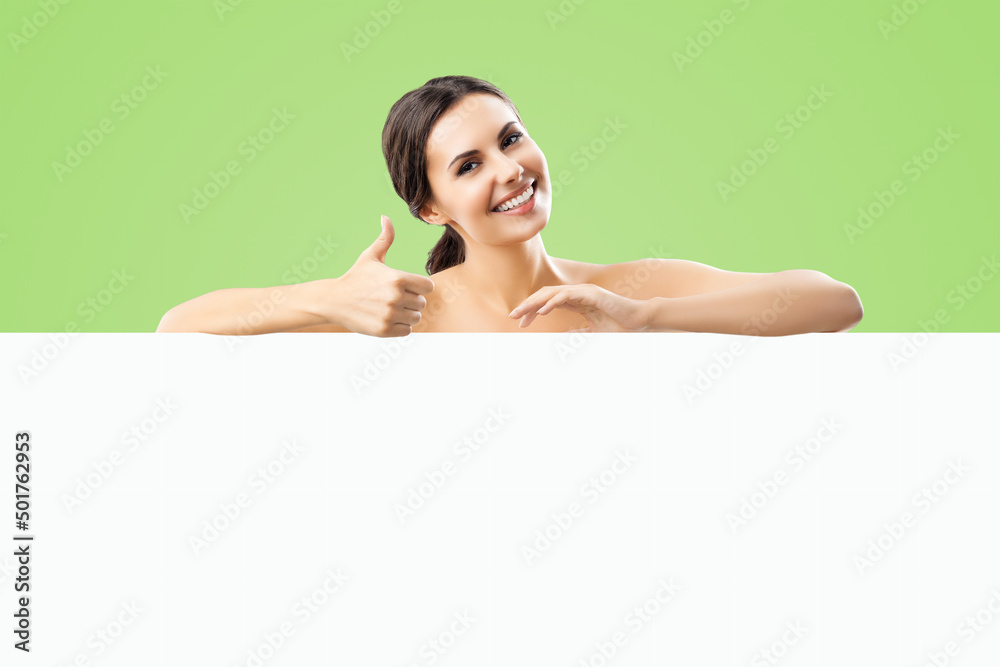 Excited smiling brunette woman with naked shoulders, undress, showing thump up hand sign gesture, standing behind white banner mock up signboard with copy space area, over green color background. Excited smiling brunette woman with naked shoulders, undress, showing thump up hand sign gesture, standing behind white banner mock up signboard with copy space area, over green color background.