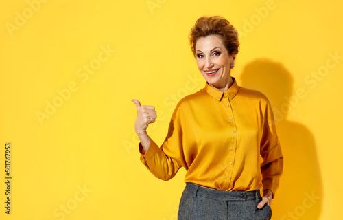 Smiling woman points aside with thumb, shows on copy space for your text. Photo of attractive elderly woman in yellow shirt on yellow background