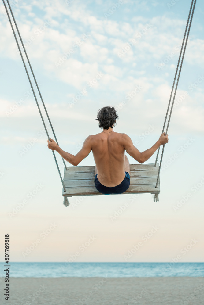 Young man traveler enjoying his summer vacation on swing by the sea. Swinging on the beach. Back view