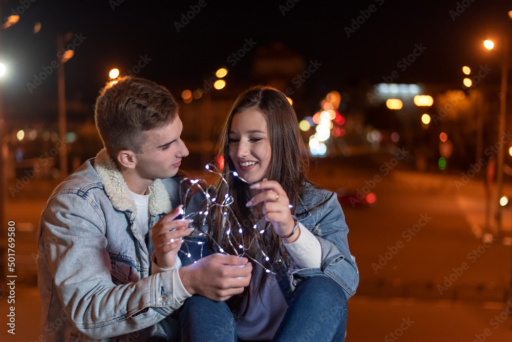 Couple of students in love are sitting on evening city background with garland in their hands.