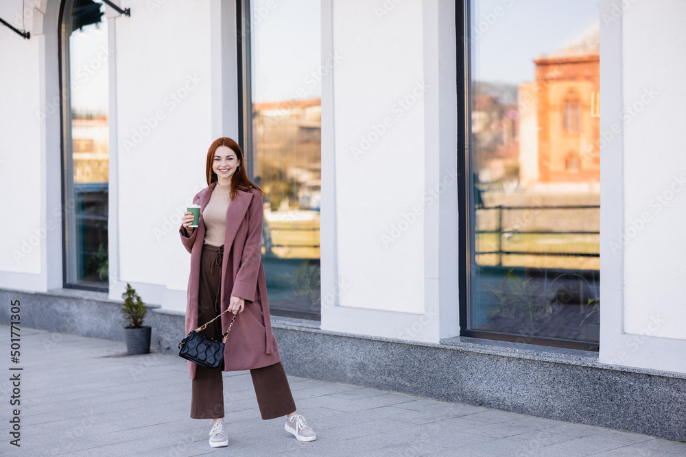full length of happy woman with handbag holding paper cup on street.