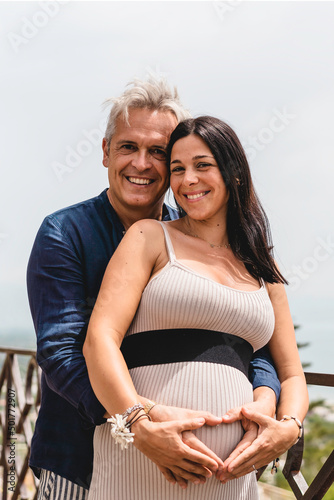 Happy couple with pregnant woman standing against a beautiful sea landscape during the summer vacation - Beautiful wife and cool middle aged man laughing together expecting a baby - Pregnancy concept