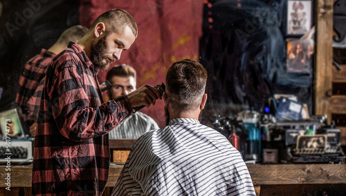 Hands of barber with hair clipper, close up. Bearded man in barbershop. Haircut concept. Man visiting hairstylist in barbershop. Barber works with hair clipper. Hipster client getting haircut