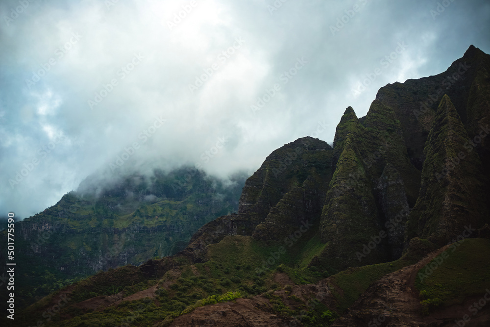 The gorgeous rugged wilderness and cliffs of Kauai's Napali Coast in Hawaii, with low clouds and mist hanging over the mountain peaks under a stormy grey sky The gorgeous rugged wilderness and cliffs of Kauai's Napali Coast in Hawaii, with low clouds and mist hanging over the mountain peaks under a stormy grey sky