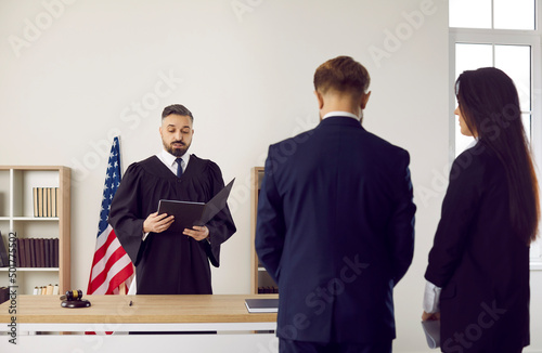 Serious fair male American judge in gown uniform enforces punishment on young man. Lawyer and defendant standing and listening to judge giving judgment, back, rear view. Court trial, law in US concept