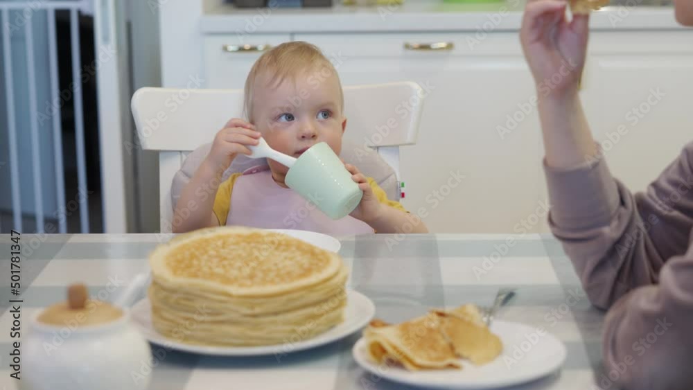 Cute kid sitting in booster seat fixed on top of dining chair, one year old baby boy with mom eating pancakes at the dinner table. High quality 4k footage Cute kid sitting in booster seat fixed on top of dining chair, one year old baby boy with mom eating pancakes at the dinner table. High quality 4k footage