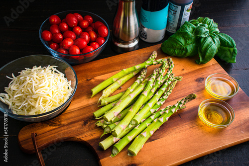 Stemmed Asparagus on a Wood Cutting Board with Other Ingredients: Prepping asparagus, tomatoes, olive oil, and shredded mozzarella cheese