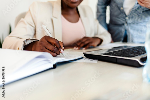 Unrecognizable business woman sitting with college in her home office and writing in her notebook. Shot of an unrecognisable woman writing in a notebook while working from home. Close up.