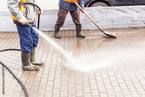 Two Man, worker in yellow orange vest and rubber boots washing sidewalk, road. Cleaning street with hose with water and shovel