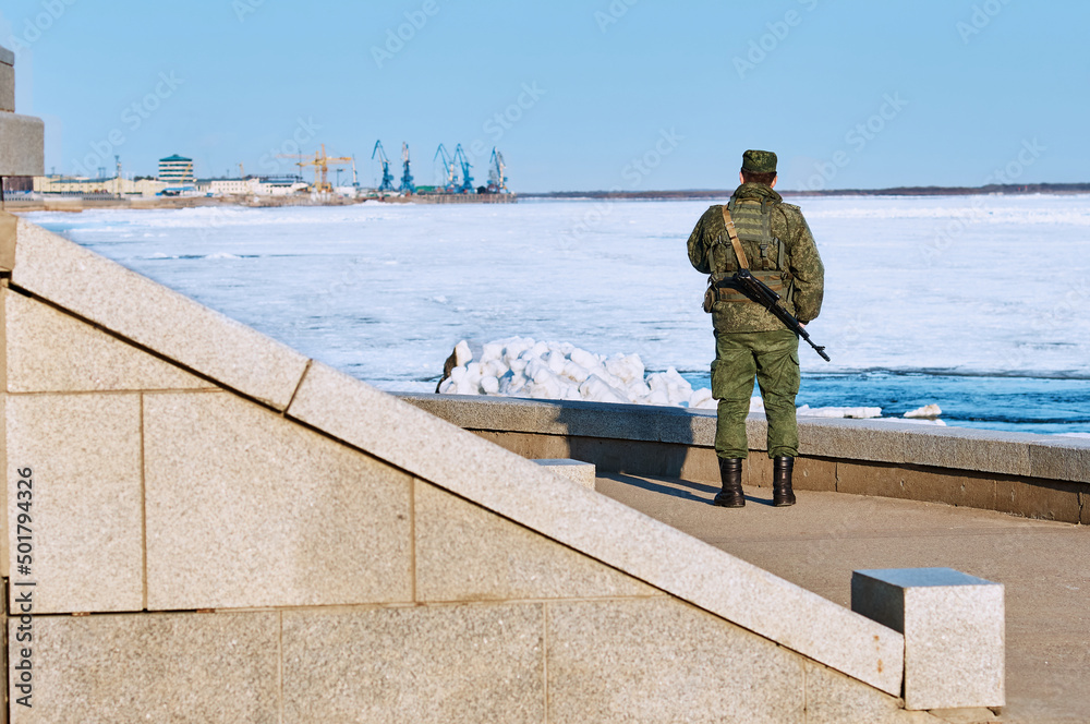 An armed Russian border guard guards the state border. View of a soldier from the back. Granite embankment of the Amur River on a spring sunny day. Before the ice drift on the river.