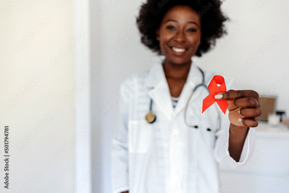 African woman wearing doctor uniform holding support red ribbon smiling at camera. Medical health care, help and hope. Sign of healthcare medicine campaign holding in female doctor. Cancer concept. African woman wearing doctor uniform holding support red ribbon smiling at camera. Medical health care, help and hope. Sign of healthcare medicine campaign holding in female doctor. Cancer concept.