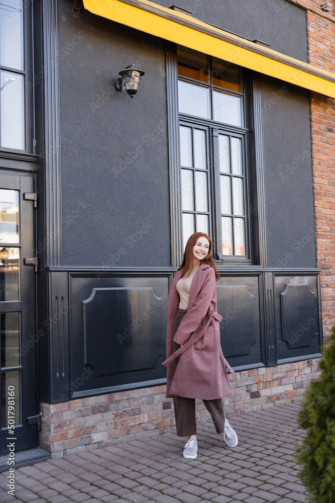 full length of happy woman in stylish coat walking near modern building.