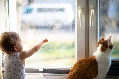 baby girl and ginger cat looking through the window at sunny day, natural dirty glass background