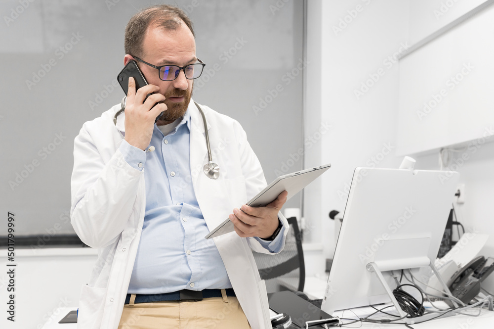 Male physician with stethoscope sitting at desk, talking on mobile phone and looking at digital tablet, discussing a patient diagnostic. High quality photography Male physician with stethoscope sitting at desk, talking on mobile phone and looking at digital tablet, discussing a patient diagnostic. High quality photography