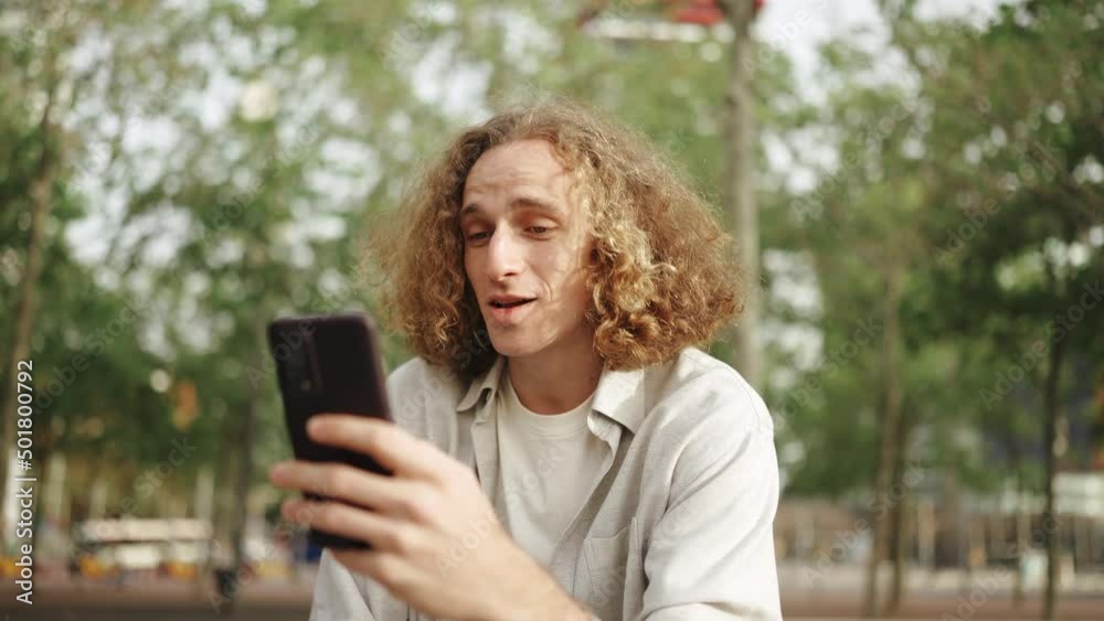 Positive curly-haired man talking by video call on phone while sitting outdoors Positive curly-haired man talking by video call on phone while sitting outdoors