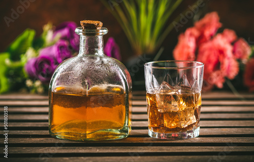 Glass of whiskey with ice and bottle with liquor on a wooden table with some color flowers. Alcoholic beverage ready to drink.