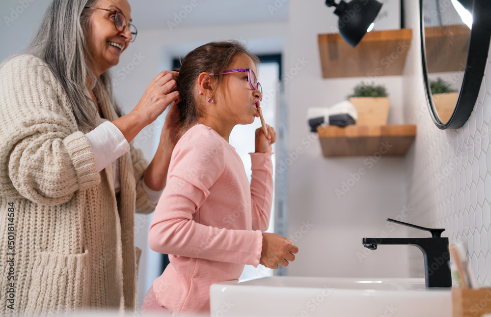 Senior grandmother making a ponytail to her granddaughter indoors in bathroom in the morning. Senior grandmother making a ponytail to her granddaughter indoors in bathroom in the morning.