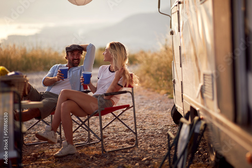 Couple at sunset cheering with drinks. Sitting in front of camper rv. Fun, togetherness, travel, nature concept.