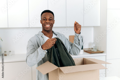 Smiling attractive black man unpacked his parcel, happy about getting a long expected order. Handsome guy shopping in internet stores, buying new clothes online, online shopping, delivery concept