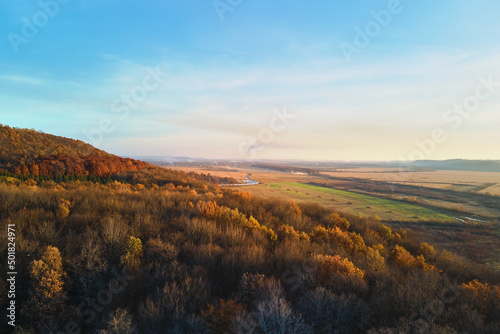 Aerial view of hills covered with dark mixed pine and lush forest with green and yellow trees canopies in autumn mountain woods at sunset. Beautiful autumnal evening landscape