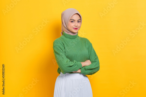Cheerful beautiful Asian woman in green sweater crossing hands and smiling at camera isolated over yellow background