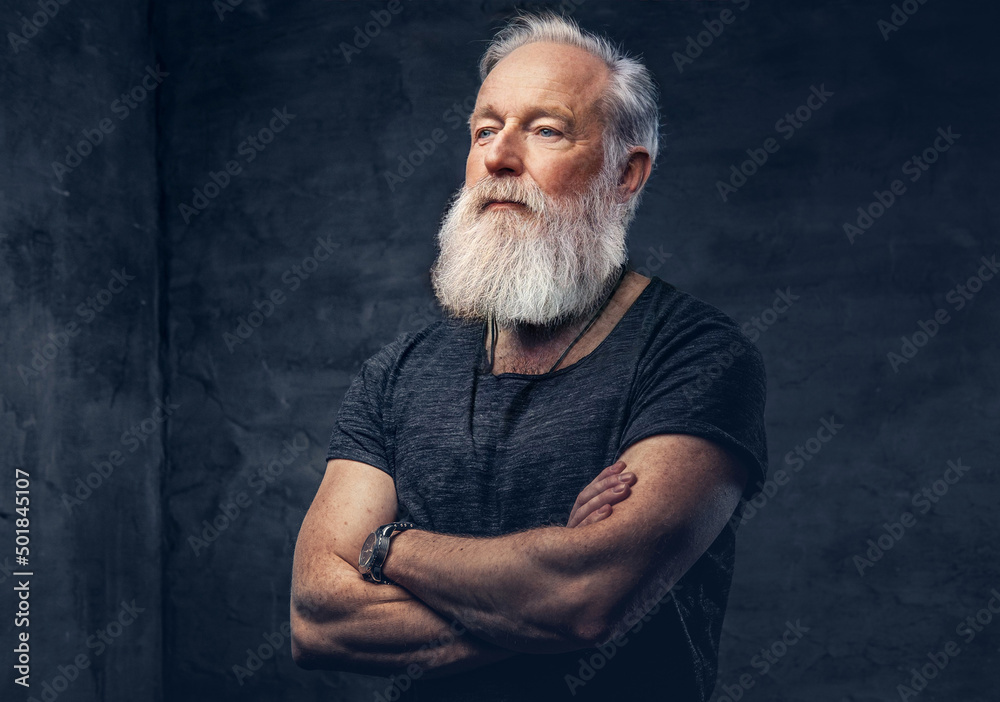 Studio shot of serious old man with muscular build and crossed arms against dark background.